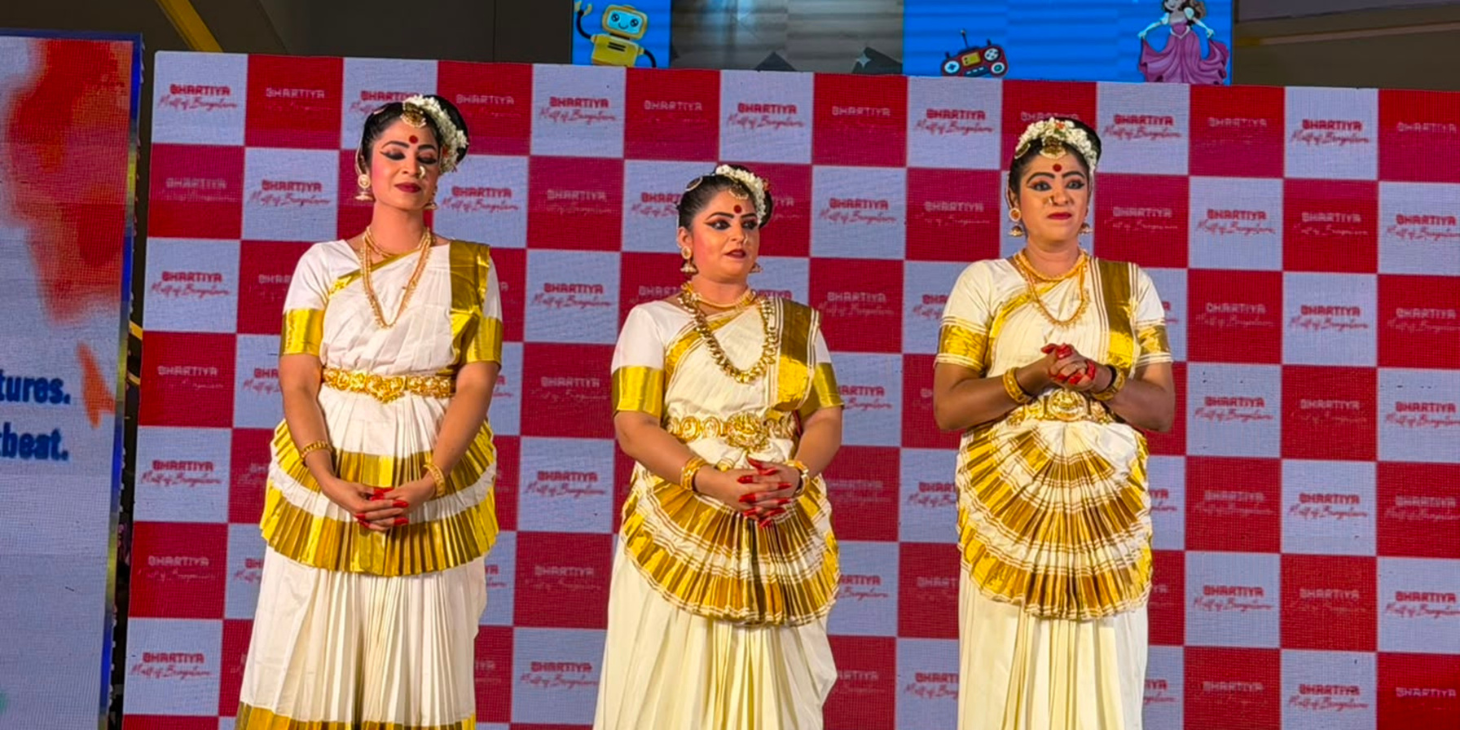 Three women in white and gold traditional Indian dance costumes with floral headpieces and gold jewelry stand on a stage in front of a red-and-white branded backdrop, posed with hands folded in front.