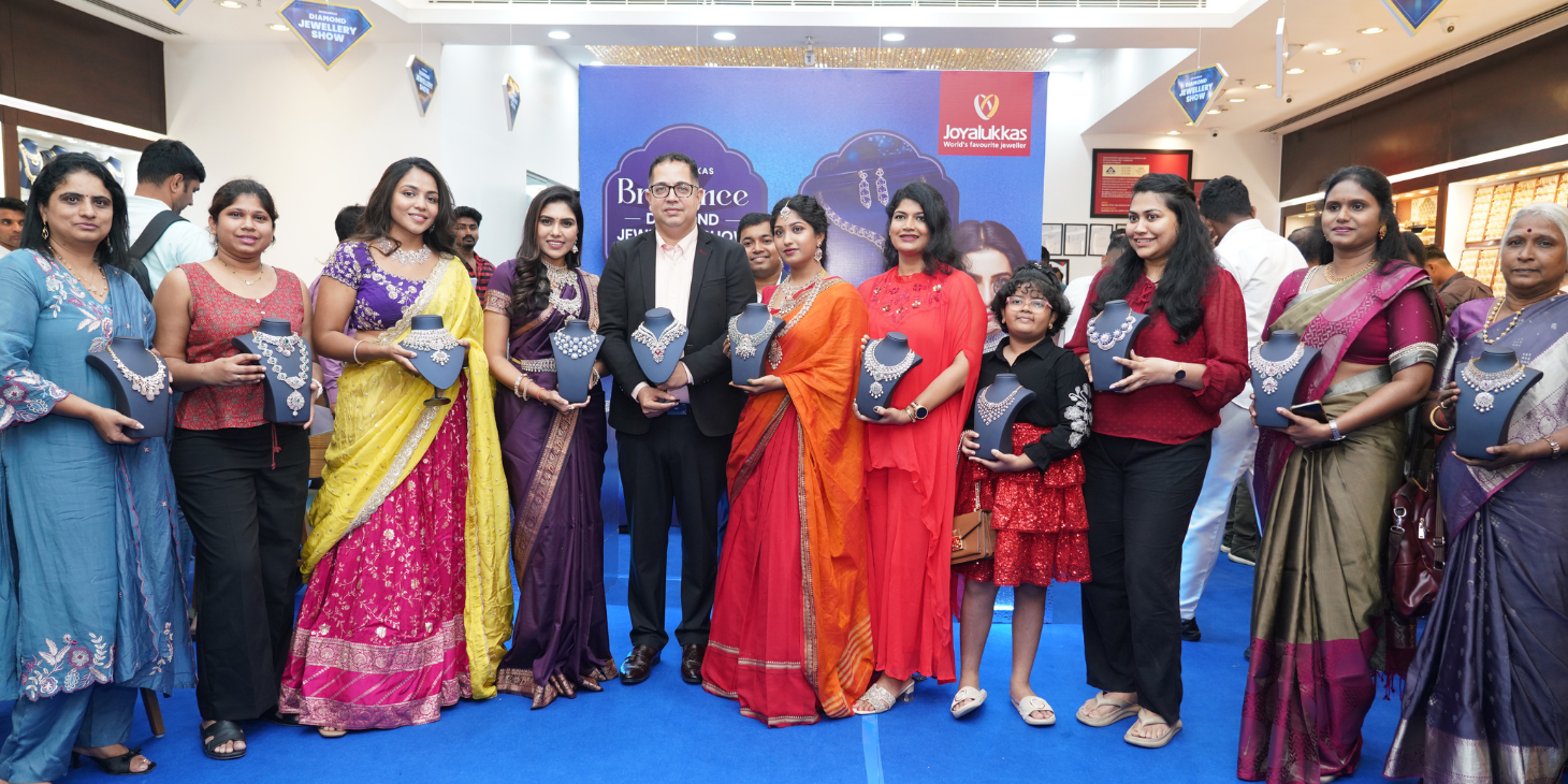 Group of men and women in colorful traditional attire posing with necklace displays at a jewelry show in a blue-carpeted hall.
