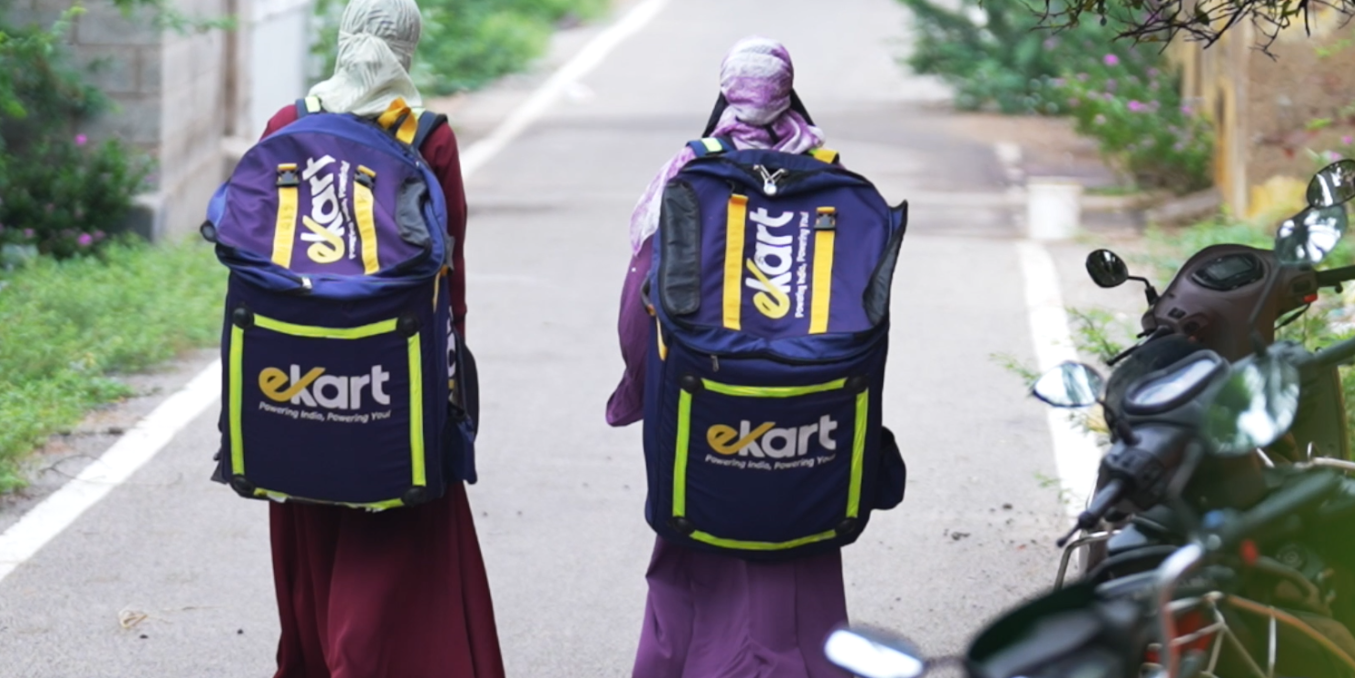 Two people with large blue eKart backpacks walk down a street, seen from behind, with motorbikes parked to the right.